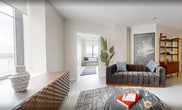 Suite 808 at The Terminal Building is a thoughtfully designed 690-square-foot apartment located in the heart of Rochester, NY. The unit features two bedrooms, one bathroom, and a combined kitchen and living area, all finished with light wood-look flooring and a neutral palette throughout. The open living room is bright and airy, with large windows offering city views, and accommodates a spacious seating area anchored by a round marble-top coffee table. The galley-style kitchen is well-appointed with dark wood cabinetry, white quartz countertops, a subway tile backsplash, stainless steel appliances including a French door refrigerator and gas range, and a deep undermount sink with a professional-style faucet. A light-filled hallway connects the bedrooms, featuring a built-in window bench and clean white millwork. The primary bedroom spans 120 square feet and includes direct access to the bathroom. The second bedroom features a charming original door with frosted glass, adding a nod to the building's historic character. The bathroom is finished with a glass-enclosed walk-in shower clad in subway tile, wood-toned cabinetry, and a white quartz vanity top.