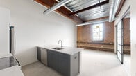 Located at 356 Hertel Ave in Buffalo's vibrant Hertel Avenue corridor, this 1-bedroom, 1-bathroom unit showcases a compelling industrial-chic aesthetic rooted in its converted building origins. The open living and kitchen area is the heart of the space, anchored by a sleek island with a stainless steel sink, light quartz countertops, and charcoal gray cabinetry. A full suite of stainless steel appliances — including a range, microwave, and refrigerator — pairs with crisp white cabinetry and a subway tile backsplash to blend modern convenience with raw character. Polished concrete floors run throughout, complementing the soaring ceilings where exposed steel beams, wood decking, and industrial ductwork command attention. Original brick walls add warmth and texture, while large factory-style windows flood the living area with natural light. The generously sized primary bedroom offers ample room for a full bedroom suite. The bathroom features classic white subway tile wainscoting, a clean vanity, and a walk-in glass-enclosed shower. A hallway connects the rooms efficiently, completing this well-appointed urban retreat that masterfully balances historic character with contemporary finishes.