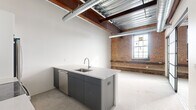 Located at 356 Hertel Ave in Buffalo's vibrant Hertel Avenue corridor, this 1-bedroom, 1-bathroom unit showcases a compelling industrial-chic aesthetic rooted in its converted building origins. The open living and kitchen area is the heart of the space, anchored by a sleek island with a stainless steel sink, light quartz countertops, and charcoal gray cabinetry. A full suite of stainless steel appliances — including a range, microwave, and refrigerator — pairs with crisp white cabinetry and a subway tile backsplash to blend modern convenience with raw character. Polished concrete floors run throughout, complementing the soaring ceilings where exposed steel beams, wood decking, and industrial ductwork command attention. Original brick walls add warmth and texture, while large factory-style windows flood the living area with natural light. The generously sized primary bedroom offers ample room for a full bedroom suite. The bathroom features classic white subway tile wainscoting, a clean vanity, and a walk-in glass-enclosed shower. A hallway connects the rooms efficiently, completing this well-appointed urban retreat that masterfully balances historic character with contemporary finishes.