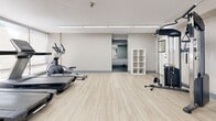 Located at 66 Mooregate Crescent in Kitchener, Ontario, this fitness facility offers a well-appointed, single-level space divided into two interconnected exercise rooms. The layout provides generous floor area designed to comfortably accommodate a wide variety of workout equipment and exercise activities.

The space features light wood-style vinyl flooring throughout, lending a clean and contemporary aesthetic that is both durable and easy to maintain. Neutral-toned walls and bright recessed and overhead lighting create an open, energizing atmosphere conducive to physical activity.

The main exercise area is lined with floor-to-ceiling windows along one wall, flooding the room with natural light and offering views of the surrounding outdoor greenery. Large wall-mounted mirrors further enhance the sense of space while allowing users to monitor their form. This room is equipped with spin bikes, strength machines, and resistance equipment, along with a medicine ball rack and a fitness reference poster.

The adjoining room, accessible through a wide interior doorway, features additional cardio equipment including elliptical trainers, as well as cable and weight machines. A storage shelf unit provides organizational space for accessories.

Together, the two rooms form a comprehensive fitness environment that is bright, functional, and thoughtfully arranged to support a full range of strength and cardio training needs.
