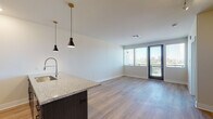 This well-appointed single-floor unit offers a thoughtfully designed open-concept layout that seamlessly connects the kitchen and living area. The kitchen serves as a central focal point, anchored by a striking dark-cabinetry island topped with speckled granite countertops and equipped with a professional-style pull-down faucet. Pendant lights with matte black shades hang above the island, adding a modern aesthetic touch. The kitchen also features white upper cabinetry, stainless steel appliances including a refrigerator with ice and water dispenser, a microwave, and a gas range. Light wood-tone flooring runs throughout the main living areas, contributing to a clean, contemporary feel. The living area is bathed in natural light courtesy of large windows and a glass door that opens to an outdoor area, offering views of the surrounding cityscape. The bedroom is generously sized, finished with plush carpeting, a ceiling fan, and a window framing outdoor views. The bathroom is elegantly appointed with dark-framed mirrors, a wide granite vanity countertop, and dark cabinetry consistent with the rest of the unit's design language. Track lighting, recessed lights, and clean white walls throughout complete the modern, polished ambiance of this urban residence.