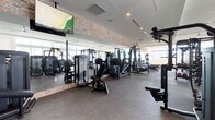 This single-floor fitness center offers a well-appointed workout environment designed to accommodate a wide range of exercise needs. The space is defined by its striking exposed brick accent wall, which adds a warm industrial character and contrasts beautifully with the clean, modern finishes throughout. Large windows allow abundant natural light to fill the room, creating an open and energizing atmosphere. A full-length mirror wall runs along one side of the gym, visually expanding the space and allowing users to monitor their form during workouts. The flooring consists of durable rubber gym matting, providing safety and comfort underfoot. The equipment lineup is extensive, featuring a variety of strength training machines including cable systems, leg press, chest press, and multi-use cable stations, all finished in sleek black. Medicine balls and additional accessories are neatly organized along the far wall. Overhead, recessed lighting and a mounted flat-screen television enhance the functionality and comfort of the space. The overall ambiance is clean, professional, and inviting — a fully functional fitness facility suited for a broad spectrum of training goals.