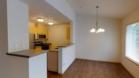 This single-floor residence is designed with an open-concept layout that connects the kitchen, dining, and living areas into one cohesive and functional space. The kitchen is equipped with stainless steel appliances, including a refrigerator, microwave, and gas range, and features warm-toned cabinetry paired with dark countertops. A peninsula with built-in shelving on the living room side serves as a natural divider between the kitchen and the adjoining spaces, offering both additional storage and a casual surface for everyday use. The dining area, defined by hardwood-style flooring, is lit by an elegant multi-arm chandelier that adds a decorative focal point to the room. The living area transitions to plush carpeting, creating a comfortable and inviting atmosphere. A hallway visible from the main living space leads to additional rooms, providing a clear and practical flow throughout the unit. The overall color palette of soft whites and warm wood tones lends the space a clean, neutral aesthetic that is well-suited for a variety of interior styles. Built-in shelving near the kitchen entry adds further storage utility to this thoughtfully arranged home.