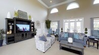 This single-floor clubhouse offers a thoughtfully designed communal space that blends elegance with comfort. The main lounge area immediately impresses with its soaring ceilings adorned with classic crown molding, creating an open and airy atmosphere. Large arched transom windows positioned high on the walls allow generous natural light to flood the interior, complementing the bright, neutral wall tones throughout.

The lounge is furnished with a curated mix of seating, including a patterned armchair, a plush blue sofa with coordinating accent pillows, and a pair of upholstered blue armchairs arranged around a dark wood coffee table — ideal for conversation and socializing. A substantial dark wood entertainment center anchors one wall, housing a flat-screen television alongside decorative shelving accented with plants and artwork.

Tall, white-trimmed French doors and large windows along the exterior wall provide views to the outdoors and connect the interior to the surrounding environment, enhancing the sense of space. Potted indoor plants are strategically placed throughout, adding warmth and a natural touch to the refined décor. The overall ambiance is inviting and polished, making this clubhouse a comfortable and attractive gathering place for residents.