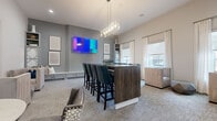 This well-appointed amenity lounge offers a thoughtfully designed social space spread across a single floor. The room is anchored by a substantial dark wood bar-height island with seating for several guests, illuminated by a contemporary rectangular chandelier that adds a touch of elegance overhead. The walls are finished in a sophisticated grey tone, providing a neutral backdrop that complements the eclectic mix of teal leather bar chairs, tufted lounge seating, and patterned accent chairs throughout.

Multiple distinct seating zones encourage different uses of the space — from casual conversation areas with plush armchairs to intimate dining nooks with round pedestal tables and mixed upholstered chairs. A built-in banquette with a crisp white surround and decorative paneling lines one wall beneath a large wall-mounted TV, offering a cozy viewing area. A second wall-mounted display serves the bar seating zone.

The space is further enlivened by a shuffleboard table positioned near large windows that flood the room with natural light through white drape curtains. Abstract artwork adorns the walls, contributing to the modern, curated aesthetic. Neutral-toned carpet grounds the room, while tall ceilings enhance the overall sense of openness and comfort throughout this versatile gathering space.