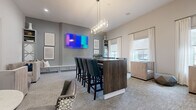 This well-appointed amenity lounge offers a thoughtfully designed social space spread across a single floor. The room is anchored by a substantial dark wood bar-height island with seating for several guests, illuminated by a contemporary rectangular chandelier that adds a touch of elegance overhead. The walls are finished in a sophisticated grey tone, providing a neutral backdrop that complements the eclectic mix of teal leather bar chairs, tufted lounge seating, and patterned accent chairs throughout.

Multiple distinct seating zones encourage different uses of the space — from casual conversation areas with plush armchairs to intimate dining nooks with round pedestal tables and mixed upholstered chairs. A built-in banquette with a crisp white surround and decorative paneling lines one wall beneath a large wall-mounted TV, offering a cozy viewing area. A second wall-mounted display serves the bar seating zone.

The space is further enlivened by a shuffleboard table positioned near large windows that flood the room with natural light through white drape curtains. Abstract artwork adorns the walls, contributing to the modern, curated aesthetic. Neutral-toned carpet grounds the room, while tall ceilings enhance the overall sense of openness and comfort throughout this versatile gathering space.
