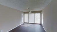 This single-floor space offers a clean, functional layout designed to maximize comfort and natural light. The main living area is a standout feature, showcasing rich, dark hardwood flooring that adds warmth and a sense of depth to the room. Large windows fitted with horizontal blinds line the walls, allowing generous amounts of natural light to filter in and create a bright, airy atmosphere throughout the day. A sliding glass door provides additional access to the outdoors while further enhancing the flow of light into the space. The ceiling features a classic textured finish, adding subtle visual interest overhead, and is equipped with a ceiling fan with an integrated light fixture — a practical touch that supports both air circulation and ambient lighting. Neutral-toned walls in a soft beige complement the flooring and contribute to the space's calm, understated aesthetic. The overall design is simple and adaptable, providing a versatile canvas suitable for a variety of furnishing styles and functional arrangements. The combination of natural light, durable flooring, and a well-considered layout makes this a welcoming and practical living environment.