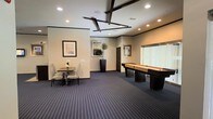 This single-floor amenity space is thoughtfully designed to offer a blend of leisure and social gathering in a polished, contemporary setting. The room is anchored by a full-length shuffleboard table, making it an ideal destination for recreation and entertainment. A dedicated dining area with a wood table and upholstered chairs provides a comfortable spot for casual meals or conversation. The interior is finished with deep blue-toned carpeting that adds a sense of warmth and sophistication to the space. Recessed ceiling lighting illuminates the room evenly, while a distinctive geometric ceiling fixture serves as a striking architectural focal point overhead. Crown molding runs along the upper walls, lending a refined, finished look to the overall aesthetic. Framed artwork is displayed throughout, adding visual interest and a curated touch to the neutral-toned walls. A tall decorative planter with lush greenery introduces a natural element to the space. Large windows allow natural light to filter in, enhancing the open and airy atmosphere. The layout flows seamlessly, offering ample room for movement and multiple activities simultaneously, making this space well-suited for both relaxed gatherings and organized events.