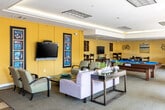 This well-appointed single-floor community clubroom offers a versatile and inviting space designed for socializing and recreation. The interior is characterized by warm yellow accent walls, recessed and tray ceiling lighting, and neutral carpeting that ties the various activity zones together seamlessly.

The lounge area serves as the centerpiece of the space, furnished with comfortable sofas, upholstered armchairs, and glass-top coffee tables adorned with decorative potted plants. A wall-mounted flat-screen television is flanked by framed artwork, creating an ideal setting for casual gatherings. Adjacent to the lounge, high-top bistro tables and bar-height chairs provide additional seating options for smaller groups.

The game area features a full-sized pool table with a vibrant blue felt surface, as well as a dedicated card or game table surrounded by dark wood chairs — ideal for organized game nights or informal play. A cozy nook with a glass-top bistro table and decorative wall art offers a quieter spot for intimate conversation.

Large sliding glass doors along one wall flood the space with natural light and open directly to an outdoor courtyard, where palm trees and architectural surroundings are visible, enhancing the bright and airy ambiance. The seamless transition between the interior and exterior adds to the overall openness and appeal of this well-designed communal amenity space.