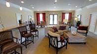 This single-story space is designed as a welcoming communal gathering area, thoughtfully arranged to accommodate both intimate conversations and larger social interactions. The open-concept layout is anchored by a generously sized seating area at its center, featuring a neutral-toned sectional sofa accented with deep red and burgundy throw pillows, complemented by dark-wood accent chairs with cross-back detailing and a woven rattan coffee table.

Recessed lighting is evenly distributed across the ceiling, which is framed by elegant crown molding, lending the space a polished, finished look. A set of French doors along the back wall allows natural light to flow freely into the interior while offering views of the outdoors. Flanking the doors are tall windows dressed in rich red curtains, adding warmth and a sense of drama to the otherwise neutral color palette of beige walls and carpeted flooring.

Toward one side of the room, a casual dining area features rattan-style chairs and a small table, providing a functional spot for more informal gatherings. Tall bar-height seating with dark wood barstools adds another distinct social zone. Decorative lamps, mirrored furniture accents, and potted plants contribute to the overall refined yet comfortable ambiance of this well-appointed space.