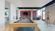 Located at 230 E Burnside St in Portland, Oregon, this single-floor amenity lounge delivers a contemporary, social atmosphere designed for recreation and gathering. The space is defined by its open layout, anchored by a solid natural wood billiards table positioned atop a woven area rug over large-format porcelain tile flooring. A striking feature wall clad in diagonal warm wood planking serves as a bold backdrop, housing a wall-mounted flat-screen television flanked by ambient uplighting that casts a rich, inviting glow.

Flanking the feature wall, sleek grey-green cabinetry with clean-lined hardware forms a functional wet bar complete with an undermount sink, providing convenient amenities for entertaining. Exposed structural concrete columns and a partially open ceiling with a dark steel grid give the space an industrial-modern character, contrasting effectively with the crisp white walls.

Floor-to-ceiling windows along one side of the lounge flood the interior with natural light and offer sweeping elevated views of the Portland cityscape. Cafe-height tables with modern stools are positioned to take full advantage of this panorama. An adjacent glass-enclosed room with colorful geometric wall art provides a secondary gathering or meeting area, adding versatility to the overall layout.