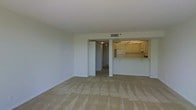 This single-floor residence offers a clean, functional layout designed for comfortable everyday living. The main living area is generously proportioned, featuring neutral-toned wall-to-wall carpeting that lends a warm and understated feel to the space. Smooth white walls and a flat ceiling with integrated HVAC vents contribute to a bright, airy atmosphere throughout.

A defining feature of the layout is the open pass-through kitchen, which connects seamlessly to the living area. The kitchen is equipped with white cabinetry, under-cabinet lighting, and standard appliances including a refrigerator. The countertop pass-through creates a natural flow between the cooking and living spaces, making the unit feel open and cohesive.

A hallway extends from the living area, leading to additional rooms within the unit, suggesting a practical and well-organized floor plan. The transition from carpeted to hard flooring in the hallway adds subtle visual contrast between zones.

Overall, the space presents a straightforward, move-in-ready environment with a neutral palette throughout, offering a versatile canvas suitable for a variety of furnishing styles and personal touches.