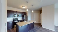 This single-floor space showcases a thoughtfully designed open-concept layout that seamlessly connects the kitchen and living areas, creating a cohesive and functional environment. The kitchen is a standout feature, highlighted by rich, dark espresso-toned cabinetry that provides ample storage and a bold aesthetic contrast against the neutral-toned walls. A large center island with granite countertops and a double-basin sink serves as both a practical workspace and a natural gathering point, illuminated by two elegant pendant lights that hang from above. Stainless steel appliances, including a refrigerator, range, and built-in microwave, add a polished, contemporary finish. The flooring throughout is a warm, dark hardwood that ties the space together with a sense of continuity and warmth. Recessed and flush-mount ceiling lighting ensures the area remains bright and inviting. The open sightlines from the kitchen extend toward what appears to be an entryway and additional rooms, suggesting a well-connected floor plan. Clean white doors and neutral wall tones contribute to an overall ambiance that feels modern, airy, and move-in ready.