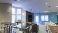 This single-floor communal lounge is designed to offer a welcoming and versatile gathering space. The room's most striking feature is a floor-to-ceiling stacked stone fireplace, which serves as a dramatic focal point and anchors the seating area. A wall-mounted flat-screen television is positioned directly above the fireplace, making it a natural entertainment hub. The vaulted ceiling adds a sense of openness and grandeur, while recessed lighting throughout ensures the space is well-illuminated at all times. Large windows along one wall flood the interior with natural light, offering views of the surrounding trees and enhancing the airy atmosphere. The layout is thoughtfully organized, with a high-top dining table and chairs in the foreground and a casual lounge area featuring upholstered seating arranged toward the fireplace. An accent wall in bold blue adds a pop of color, contrasting with the neutral tones of the walls and flooring. Glass-paneled doors provide additional light and access to an adjoining area, further contributing to the open, connected feel of this well-appointed communal space.