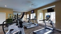 Located at 7331 Skyline Dr in Columbus, Ohio, this single-floor fitness center is designed to offer a well-equipped and comfortable workout environment. The space features a robust selection of cardio machines, including multiple treadmills and elliptical trainers from True Fitness, arranged in an open layout that promotes ease of movement and accessibility. Large wall-mounted mirrors line one side of the room, visually expanding the space and allowing users to monitor their form during exercise. Overhead, a ceiling fan supports ventilation and comfort, while recessed lighting ensures the room is well-lit throughout. Multiple windows along the walls invite an abundance of natural light and provide views of the surrounding greenery, creating a bright and energizing atmosphere. A wall-mounted flat-screen television is positioned centrally, offering entertainment during workouts. The flooring features a durable, patterned surface suited for a high-traffic fitness environment. The warm-toned walls contribute to an inviting ambiance, making this fitness center a functional and welcoming space for residents and users seeking a convenient on-site exercise facility.