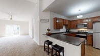 This single-floor residence offers a thoughtfully designed open layout that promotes a seamless flow between living and dining areas. The kitchen is a standout feature, outfitted with rich medium-toned wood cabinetry, dark granite-style countertops, and a full suite of stainless steel appliances including a range, microwave, and refrigerator. A peninsula with a breakfast bar provides additional counter space and casual seating with two upholstered bar stools. Tile flooring in the kitchen adds durability and easy maintenance. A sliding barn door adds a modern architectural touch near the kitchen entry.

The living area benefits from soaring vaulted ceilings and a ceiling fan, creating an open, airy atmosphere. Plush wall-to-wall carpeting runs throughout the living and bedroom spaces, adding warmth underfoot. A sliding glass door in the living area brings in natural light and provides access to the outdoors. The bedrooms are generously proportioned, each featuring carpeted floors and windows with blinds that let in natural light while maintaining privacy. The bathroom is clean and functional, featuring tile flooring, a combined tub and shower with a curtain, a vanity with wood cabinetry, and crisp white fixtures throughout.