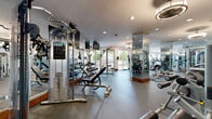 This fitness center is designed to accommodate a comprehensive range of workout routines within a thoughtfully organized, single-floor layout. The space is divided into distinct functional zones, each tailored to specific training needs. The cardio area features a row of modern treadmills and elliptical machines aligned along large windows that overlook greenery outside, filling the room with natural light and creating an open, airy atmosphere. Decorative drum-shade ceiling fixtures complement the recessed lighting throughout, contributing to a polished, contemporary aesthetic.

The strength training zone is well-appointed with Precor cable machines, a multi-station cable crossover unit, a full dumbbell rack, adjustable benches, and a variety of selectorized machines targeting major muscle groups. A dedicated free weights area includes barbells, weight plates, and open floor space with rubber mat flooring for functional movements. Floor-to-ceiling mirrors are strategically positioned throughout, visually expanding the space and allowing users to monitor their form. A stability ball and medicine ball rack round out the functional fitness offerings. The durable rubber flooring provides both safety and comfort underfoot across all training areas.