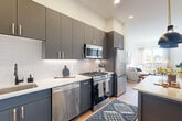 This well-designed one-bedroom, one-bathroom residence in Everett, MA offers a thoughtfully laid-out single-floor plan with a clean, modern aesthetic throughout. Light hardwood flooring flows seamlessly across the entire space, complementing the bright white walls and recessed lighting overhead.

The kitchen is a standout feature, showcasing sleek charcoal-gray flat-front cabinetry extending to near ceiling height, paired with crisp white quartz countertops and a white geometric tile backsplash. A full suite of stainless steel appliances — including a gas range, built-in microwave, dishwasher, and refrigerator — makes this an exceptionally functional space for cooking and entertaining. The kitchen opens directly into the living area, creating an airy, connected feel.

The living room benefits from large windows that invite generous natural light and offer open exterior views, creating a bright and welcoming atmosphere. The bedroom is a comfortable, well-proportioned retreat featuring its own large window with outdoor views, a ceiling fan, and ample natural light. Three closets throughout the unit provide notable storage capacity.

The bathroom is finished with white stacked subway tile in a walk-in shower enclosed by a sleek black-framed glass door, paired with light wood flooring for a spa-like, contemporary feel.