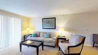 This single-floor suite offers a thoughtfully designed layout that balances comfort and functionality across several distinct areas. The living room serves as a welcoming focal point, furnished with a plush beige sofa accented by teal throw pillows, a coordinating armchair, and a dark wood coffee table. Warm lamp lighting and framed wall art contribute to an inviting, relaxed atmosphere, while a sliding glass door with vertical blinds opens to an outdoor view, filling the space with natural light. A dedicated media area features a flat-screen TV and storage console, with an additional sliding door providing further outdoor access. The fully equipped kitchen is outfitted with stainless steel appliances — including a refrigerator, dishwasher, electric range, and over-range microwave — complemented by light wood cabinetry, granite countertops, and tile flooring. A curved breakfast bar with pendant lighting adds a casual dining option. The bedroom is comfortably sized, featuring a dark wood bed frame and matching nightstands, with a window that brings in natural light and views of surrounding greenery. The bathroom continues the granite countertop aesthetic, paired with a tiled floor and tub/shower combination, completing this well-rounded suite.