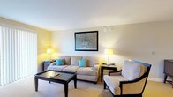 This single-floor suite offers a thoughtfully designed layout that balances comfort and functionality across several distinct areas. The living room serves as a welcoming focal point, furnished with a plush beige sofa accented by teal throw pillows, a coordinating armchair, and a dark wood coffee table. Warm lamp lighting and framed wall art contribute to an inviting, relaxed atmosphere, while a sliding glass door with vertical blinds opens to an outdoor view, filling the space with natural light. A dedicated media area features a flat-screen TV and storage console, with an additional sliding door providing further outdoor access. The fully equipped kitchen is outfitted with stainless steel appliances — including a refrigerator, dishwasher, electric range, and over-range microwave — complemented by light wood cabinetry, granite countertops, and tile flooring. A curved breakfast bar with pendant lighting adds a casual dining option. The bedroom is comfortably sized, featuring a dark wood bed frame and matching nightstands, with a window that brings in natural light and views of surrounding greenery. The bathroom continues the granite countertop aesthetic, paired with a tiled floor and tub/shower combination, completing this well-rounded suite.