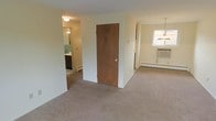 This single-floor residence offers a straightforward and functional layout with a warm, understated aesthetic. The main living area features neutral wall-to-wall carpeting throughout, creating a cohesive and comfortable feel underfoot. Soft, cream-colored walls provide a clean backdrop that lends itself well to a variety of interior styles.

A solid wood interior door separates the living spaces, offering privacy between rooms. The adjoining room to the right benefits from natural light streaming through a window, and is equipped with a wall-mounted air conditioning unit and a baseboard heater, ensuring year-round climate comfort. An elegant pendant light fixture adds a subtle decorative touch overhead.

Visible through the open doorway to the left is the bathroom, which features distinctive teal green tile work that adds a pop of color and character. A vanity with dark cabinetry is also visible, providing practical storage. The bathroom benefits from its own lighting and maintains the home's clean, utilitarian design language.

Overall, this residence presents a tidy, well-maintained environment with consistent finishes across its single floor, making it a practical and livable space suited for comfortable day-to-day living.