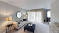 This single-floor residence presents a cohesive, neutral color palette across all rooms, creating a calm and welcoming atmosphere. The living room is a standout space, featuring soft gray walls, plush carpeting, and a wide sliding glass door fitted with vertical blinds that floods the area with natural light and provides outdoor views. A tufted navy ottoman and a classic sofa anchor the room, while a decorative mirror adds visual depth.

Adjacent to the living area, the dining space accommodates a round glass-top table beneath a pendant light fixture. Natural light enters through a window dressed with dark curtain panels, and a decorative mirror on the wall adds an elegant touch.

The kitchen is functional and efficiently laid out, featuring light wood cabinetry, dark granite-style countertops, a stainless steel sink, and white appliances including a gas range and dishwasher.

The bedroom offers a restful retreat with soft gray-blue walls, plush carpet, and a window allowing ample natural light. A white dresser with mirror provides generous storage.

The bathroom includes a tub-shower combination with tile surround, complemented by warm neutral tones and decorative wall accents, completing this well-appointed home.