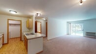 This single-floor residence offers a practical and well-organized layout with a clean, neutral aesthetic throughout. The open-concept design connects the kitchen directly to the living area, creating a cohesive and functional space. The kitchen features warm oak cabinetry lining both walls in a galley-style arrangement, white appliances including a range, refrigerator, and dishwasher, a double stainless steel sink, and light hardwood-style flooring. A peninsula with a secondary sink separates the kitchen from the main living area, providing additional counter space and a natural transition between zones.

The spacious living area is finished with plush wall-to-wall carpeting, neutral white walls, and warm wood baseboards. Track lighting illuminates the space, and a sliding glass door opens to an outdoor area, allowing natural light to flow in and offering views of surrounding greenery. A wall-mounted HVAC unit provides climate control.

The bedroom is generously proportioned, featuring carpeted floors, a large window with natural light, a baseboard heater, and a roomy walk-in closet with built-in shelving. The full bathroom includes an oak vanity with a wide white countertop, a large mirror, and a shower-tub combo. A dedicated in-unit laundry area houses a full-sized washer and dryer with overhead shelving for added storage convenience.