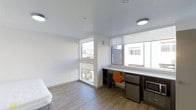 This efficiently designed studio with one bathroom offers a smart, modern layout that maximizes every square foot. The interior features light wood-tone flooring and crisp white walls and ceilings, creating a clean and airy atmosphere throughout. Large windows along two walls flood the space with abundant natural light while framing views of the surrounding urban neighborhood, enhanced by roller shades for privacy and light control. A built-in desk and shelving unit runs beneath the windows, providing a dedicated workspace complete with a mini refrigerator and microwave — all seamlessly integrated into the design. The sleeping area is positioned to take full advantage of the natural light, contributing to an open, comfortable feel despite the compact footprint. Recessed and flush-mounted ceiling lighting supplements the natural light, ensuring the space remains bright throughout the day and evening. The overall design aesthetic is minimalist and functional, with neutral tones and contemporary finishes that lend a cohesive, well-considered look. This studio is well-suited for those seeking a practical, self-contained living space with thoughtful built-in amenities in an urban setting.