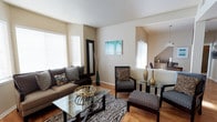This single-floor space presents a thoughtfully designed interior that balances comfort and contemporary style. The living area serves as the focal point of the space, showcasing warm hardwood flooring that flows seamlessly throughout. Neutral beige and taupe wall tones create a calm, inviting backdrop that complements the bold, patterned furnishings, including a zebra-print area rug, animal-print accent chairs, and a sleek glass-top coffee table with dark wood framing. A plush sofa dressed in decorative pillows anchors the seating arrangement, while a matching ottoman and side table add functional elegance. Decorative accents — including gold metallic vases, a framed floor mirror with a dark border, and a vibrant teal abstract wall painting — inject personality and visual interest into the room. Large windows fitted with white horizontal blinds allow ample natural light to fill the space, enhancing its open and airy feel. A glimpse into an adjoining area reveals additional living or dining space, suggesting a well-connected, flowing floor plan. The overall ambiance is warm, curated, and inviting, making it a refined environment suited for both relaxing and entertaining.