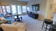 This single-floor space offers a welcoming and well-appointed living area designed for both comfort and functionality. The living room is the centerpiece of the home, featuring plush wall-to-wall carpeting that adds warmth underfoot. Large windows with white shuttered blinds flood the room with natural light, while a set of glass-paned doors provides access to an outdoor area and further enhances the bright, airy atmosphere. The neutral, warm-toned walls create a clean and versatile backdrop throughout the space. A wall-mounted flat-screen television serves as a focal point, complemented by a dark wood media console with decorative panel detailing and ample storage. Furnishings include a classic striped armchair and a dark wood coffee table, contributing to a timeless and cohesive aesthetic. Decorative accents, including a potted plant and tasteful tabletop pieces, add personality and a lived-in charm. A ceiling fan with a light fixture provides both illumination and climate comfort. The overall ambiance is calm and inviting, blending practical design with a relaxed residential character that makes the space feel immediately at home.