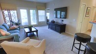This single-floor space offers a welcoming and well-appointed living area designed for both comfort and functionality. The living room is the centerpiece of the home, featuring plush wall-to-wall carpeting that adds warmth underfoot. Large windows with white shuttered blinds flood the room with natural light, while a set of glass-paned doors provides access to an outdoor area and further enhances the bright, airy atmosphere. The neutral, warm-toned walls create a clean and versatile backdrop throughout the space. A wall-mounted flat-screen television serves as a focal point, complemented by a dark wood media console with decorative panel detailing and ample storage. Furnishings include a classic striped armchair and a dark wood coffee table, contributing to a timeless and cohesive aesthetic. Decorative accents, including a potted plant and tasteful tabletop pieces, add personality and a lived-in charm. A ceiling fan with a light fixture provides both illumination and climate comfort. The overall ambiance is calm and inviting, blending practical design with a relaxed residential character that makes the space feel immediately at home.