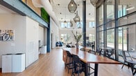 Located at 1250 Water Tower Circle in Greenville, SC, this single-floor amenity space showcases a striking contemporary design defined by soaring double-height ceilings, exposed ductwork, and bold structural steel columns that lend an industrial-modern aesthetic throughout. Wood-look flooring flows seamlessly across the open-concept layout, unifying the various functional zones within the space.

The lounge area is anchored by a vibrant blue velvet sofa, abstract wall art, and a seating arrangement bathed in natural light from oversized windows that frame lush outdoor greenery. Artistic woven pendant lights hang dramatically from above, adding sculptural character to the social gathering zones.

A dedicated dining and co-working area features a substantial live-edge communal table with bar-height seating, complemented by additional café-style round tables and chairs positioned against a dramatic floor-to-ceiling glass curtain wall that connects the interior to an outdoor courtyard, flooding the space with daylight.

The recreation zone includes a full ping-pong table and a shuffleboard table, ideal for casual entertainment. A living green moss wall installation and warm wood accents provide natural contrast to the white walls and industrial elements. The grand entry foyer features a staircase with sleek metal railings and arched glass doors, setting a welcoming and polished first impression.