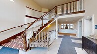 This distinguished space makes an immediate impression through its grand entrance foyer, where soaring double-height ceilings and large transom windows flood the area with natural light. A dramatic curved staircase serves as the architectural centerpiece, featuring rich hardwood treads, crisp white spindles, and dark wood handrails that gracefully ascend to an open upper-level balcony. Large-format tile flooring in the entry transitions seamlessly into plush patterned carpeting throughout the common areas, adding warmth and texture underfoot. An elegantly upholstered accent bench nestled near the staircase and a dark wood console cabinet provide refined furnishing details that complement the traditional aesthetic. Beyond the foyer, a bright and spacious gathering room opens up, highlighted by an impressive arched window arrangement that draws in abundant natural light and frames views of the surrounding exterior. A cozy seating area with a round dark wood table and leather armchairs sits beneath tall, grid-paned windows with wainscoting panels below, creating an intimate setting within the larger space. The 3D floorplan reveals an expansive, thoughtfully organized layout with extended corridor wings and interconnected common areas. Throughout, neutral warm tones, recessed lighting, and classic millwork details combine to create an atmosphere that is both elegant and inviting.