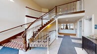 This distinguished space makes an immediate impression through its grand entrance foyer, where soaring double-height ceilings and large transom windows flood the area with natural light. A dramatic curved staircase serves as the architectural centerpiece, featuring rich hardwood treads, crisp white spindles, and dark wood handrails that gracefully ascend to an open upper-level balcony. Large-format tile flooring in the entry transitions seamlessly into plush patterned carpeting throughout the common areas, adding warmth and texture underfoot. An elegantly upholstered accent bench nestled near the staircase and a dark wood console cabinet provide refined furnishing details that complement the traditional aesthetic. Beyond the foyer, a bright and spacious gathering room opens up, highlighted by an impressive arched window arrangement that draws in abundant natural light and frames views of the surrounding exterior. A cozy seating area with a round dark wood table and leather armchairs sits beneath tall, grid-paned windows with wainscoting panels below, creating an intimate setting within the larger space. The 3D floorplan reveals an expansive, thoughtfully organized layout with extended corridor wings and interconnected common areas. Throughout, neutral warm tones, recessed lighting, and classic millwork details combine to create an atmosphere that is both elegant and inviting.