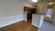 This single-floor residence offers a well-designed, open-concept layout that seamlessly connects the kitchen and living areas, creating a cohesive and functional living environment. The kitchen is equipped with warm-toned wooden cabinetry that provides ample storage, complemented by a full suite of black appliances including a range, microwave, and refrigerator. A peninsula counter with a textured finish extends into the living space, offering a natural divide between the kitchen and the adjoining area while also serving as a convenient spot for casual dining or meal prep. The flooring throughout the main living area features rich, dark hardwood-style planks that add warmth and visual depth to the space. Neutral-toned walls in a soft gray provide a clean, versatile backdrop that suits a variety of décor styles. Bright flush-mount ceiling lighting ensures the space remains well-lit throughout. The transition from the hard flooring to carpeting at the edge of the frame hints at a comfortable adjoining area, likely a living or dining space. The overall ambiance is practical and inviting, with a layout that maximizes usability across the single floor.