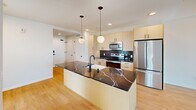 Unit #1102 at Bakers Place is a thoughtfully designed 750-square-foot, 2-bedroom, 2-bathroom condominium located in Madison, WI. The open-concept layout features light hardwood flooring throughout the main living areas, accented by warm exposed wood ceiling beams that add architectural character. The contemporary kitchen is a standout, boasting light wood cabinetry, stainless steel appliances, a dark tile backsplash, and a striking oversized island with a dark marble-style countertop and pendant globe lighting. Both bedrooms are carpeted and bathed in natural light through large black-framed windows offering elevated views of the Madison cityscape. The primary bedroom spans 160 square feet and features a sliding glass door with panoramic views. Both bathrooms are finished with sleek dark vertical tile, light wood vanities, and dark countertops — one featuring a walk-in shower and the other a full tub-shower combo. A hallway connects the unit's rooms efficiently. The overall aesthetic is clean and modern, with a neutral palette, quality finishes, and an abundance of natural light throughout.