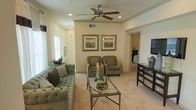 This single-floor residence offers a thoughtfully designed interior that balances comfort and style. The living room serves as the heart of the space, featuring plush, tufted upholstered seating in soft blue-gray tones complemented by coordinating accent chairs. A glass-top coffee table anchors the seating area, while a ceiling fan with a brushed nickel finish and recessed lighting provide both functionality and a polished look overhead. The walls are finished in warm neutral tones, creating a bright and inviting ambiance throughout. A pair of framed landscape artworks adds a decorative focal point above the seating area. Light-colored carpeting extends across the floor, enhancing the room's warm, welcoming feel. Natural light filters in through window treatments on the side wall, further brightening the interior. A mirrored accent dresser along one wall adds a touch of elegance and reflects light throughout the space. An open archway leads seamlessly to an adjacent room, suggesting a well-connected floor plan designed for easy flow between living areas. Overall, the space presents a refined, move-in-ready aesthetic with attention to cohesive décor and comfortable livability.