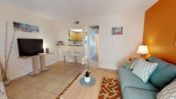 This single-floor residence offers a bright and welcoming layout with a distinctive coastal-inspired design aesthetic throughout. The living area serves as the heart of the home, highlighted by a bold orange accent wall, light tile flooring, and a teal sofa paired with a geometric-patterned area rug. Natural light filters in through large windows dressed with chevron-print curtains, creating a warm and inviting ambiance. A dedicated media corner provides additional functionality to the space.

The kitchen is efficiently designed with warm wood cabinetry, granite-style countertops, and a stainless steel sink. A raised breakfast bar with white bar stools offers a casual dining option and connects the kitchen seamlessly to the living area, enhancing the open feel of the space.

The bedroom features a soothing teal accent wall adorned with coastal artwork, tile flooring, and a window with sheer curtains that allow soft natural light to fill the room. A closet and direct access to the bathroom add practical convenience.

The bathroom is well-equipped with a wide vanity topped with granite-style countertops, rich wood cabinetry, a large mirror with overhead lighting, and a shower-tub combination. The consistent coastal theme ties the entire residence together with a relaxed, cohesive charm.