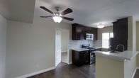 This single-floor space showcases an open kitchen and living area designed with a cohesive, modern aesthetic. The kitchen is outfitted with rich, dark espresso-toned cabinetry that extends to the upper and lower sections, providing ample storage. Light-colored countertops offer a clean contrast against the darker cabinet tones, while a white subway tile backsplash adds texture and visual interest to the cooking area. Stainless steel appliances, including a gas range and an over-the-range microwave, complete the functional kitchen setup. The flooring throughout features dark hardwood-style planks that add warmth and a polished finish to the space. A ceiling fan with a light fixture is centrally positioned, contributing to both air circulation and ambient lighting. Natural light filters in through a window near the kitchen, brightening the area. An open doorway leads to an adjacent room with light-toned carpeting, suggesting a bedroom or secondary living space. Clean white trim and baseboards frame the neutral-toned walls throughout, lending the space a tidy and well-finished appearance overall.