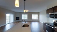 This single-floor residence offers a thoughtfully designed open-concept layout that seamlessly connects the kitchen and living areas. The kitchen serves as the heart of the space, showcasing a central island with a light stone countertop and an undermount single-basin sink paired with a curved gooseneck faucet. Stylish pendant lights hang above the island, providing warm, focused illumination that complements the overall aesthetic. The open floor plan flows effortlessly into the living area, where rich hardwood flooring extends throughout, adding warmth and a timeless appeal. Neutral-toned walls create a clean, versatile backdrop that suits a variety of interior styles. The living space is anchored by a sleek, built-in fireplace, serving as a natural focal point for the room. Two generously sized windows allow an abundance of natural light to fill the space, creating a bright and welcoming ambiance while offering views of the surrounding greenery. A flush-mount ceiling light further enhances the room's illumination. The combination of modern finishes, an open layout, and carefully selected lighting fixtures gives this single-floor home a functional yet inviting character that balances everyday practicality with contemporary style.