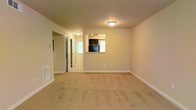 This single-floor space offers a straightforward and functional layout designed for comfortable everyday living. The main living area is generously sized, featuring wall-to-wall carpet flooring in a neutral beige tone that adds warmth underfoot. Walls are finished in a soft, neutral palette, contributing to a clean and understated aesthetic throughout.

A notable architectural detail is the pass-through window connecting the living area to the kitchen, allowing visibility and an open feel between the two spaces. The kitchen, visible through the pass-through, features dark cabinetry and appears to include standard appliances, with a window that allows natural light to filter through. Flush-mount ceiling light fixtures provide consistent overhead lighting across the living area and hallway.

A hallway extends from the living space, leading to additional rooms and featuring a door with access to natural light, suggesting an entry or exterior-facing door. A floor vent is visible along the baseboard, indicating a central HVAC system. White baseboards frame the perimeter of the rooms, providing clean transitions between the walls and flooring. Overall, the space presents a practical and move-in-ready environment with a cohesive, neutral design scheme.