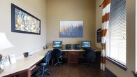 This business center is a compact yet fully functional workspace designed to meet essential professional needs. The room features an L-shaped built-in desk along the walls, providing ample surface area for multiple users simultaneously. Three computer workstations are set up side by side, each equipped with a monitor, with printers conveniently positioned between them for shared access. Ergonomic black office chairs accompany each station, ensuring comfortable use during extended sessions.

The space is finished with warm beige walls that create a welcoming, focused atmosphere. Dark hardwood flooring adds a refined, professional touch that contrasts nicely with the light-toned desk surfaces. A large, vibrant canvas painting featuring blue and gold floral tones serves as a central decorative focal point above the workstations, while a second framed artwork adds visual interest to the side wall.

Natural light filters in through a window dressed with bold orange-and-white striped curtains, brightening the room without overwhelming the workspace. A posted Business Center Rules sign near the entrance ensures orderly and considerate use of the shared amenities. Overall, the space balances functionality with a polished, inviting aesthetic.