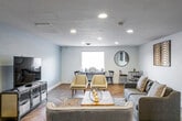 This thoughtfully designed single-floor space offers a welcoming and eclectic atmosphere, anchored by striking herringbone-patterned hardwood flooring that runs throughout the open layout. The main living area is generously proportioned, accommodating multiple seating arrangements with ease. A gray mid-century modern sofa and a curved cream accent sofa are centered around a rustic live-edge wood coffee table, creating an inviting focal point. A bold leopard-print area rug adds a layer of texture and visual interest beneath the furnishings.

Recessed lighting is installed throughout the ceiling, providing even, adjustable illumination across the space. Decorative wall art featuring geometric, wave-pattern panels adds warmth and dimension to the crisp white walls. A large round mirror with a black frame enhances the sense of depth in the room, while a compact black metal side table and mixed seating in one corner offer an intimate nook for conversation or reading.

On the opposite end of the main area, soft blue-gray walls set a calm tone around a media console with industrial-style cabinetry housing a flat-screen television. An additional dining or activity area with chairs and a side table is positioned near a window, allowing natural light to filter in. The overall ambiance is modern yet approachable, blending contemporary and eclectic design elements seamlessly.