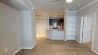 This single-floor residence showcases a thoughtfully designed open-concept layout that promotes a seamless flow between living and kitchen areas. The space is finished with warm hardwood-style flooring that runs throughout, adding a sense of continuity and comfort underfoot.

The kitchen serves as a functional centerpiece, featuring dark cabinetry contrasted by a light stone countertop with a peninsula that doubles as a breakfast bar. A classic subway tile backsplash adds texture and visual interest, while stainless steel appliances — including a range, microwave, and refrigerator — provide a modern, polished finish. Track lighting illuminates both the kitchen and the adjacent living area, ensuring bright and even coverage throughout.

Open shelving built into the wall offers practical display and storage options, enhancing the home's contemporary aesthetic. Decorative column details and crisp white trim and baseboards frame the space elegantly, contributing to its clean, refined character. A ceiling fan adds to the comfort of the living area, keeping the space well-ventilated. The overall ambiance is fresh and modern, with a neutral color palette that creates an inviting and versatile environment ready to accommodate a variety of furnishing styles.