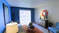 This single-story space in Amarillo, Texas, offers a comfortable and well-appointed living area designed with both style and function in mind. The living room serves as the focal point of the space, showcasing a striking deep blue accent wall that adds a bold, modern contrast to the otherwise neutral color palette of soft grays and whites throughout.

A large sliding glass door dominates the accent wall, allowing generous amounts of natural light to flood the interior while providing direct access to an outdoor area. Vertical blinds offer adjustable light control and privacy. The carpeted flooring, paired with a warm-toned area rug, adds texture and coziness underfoot.

The room is thoughtfully laid out to accommodate a seating arrangement centered around a sleek, dark coffee table, creating an inviting gathering space. A coordinating credenza along the adjacent wall provides additional storage and display space, topped with decorative accessories including a statement sunburst mirror that adds a touch of artistic character. The textured ceiling completes the interior's classic residential aesthetic, contributing to the overall warmth and livability of the space.