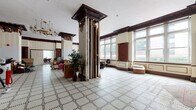 Located at 101 SE First St in downtown Evansville, Indiana, this single-floor historic commercial space showcases exceptional Beaux-Arts architectural character throughout its expansive open interior. The main hall is defined by striking structural columns adorned with elaborately carved dark wood capitals and cornices, which complement the richly detailed crown molding that runs continuously along the upper walls. Intricate geometric mosaic tile flooring in a black-and-white pattern covers the entire floor, adding a refined, classical elegance to the space. Tall, multi-pane windows line the perimeter, flooding the interior with abundant natural light and offering views of the surrounding streetscape. The high ceilings amplify the grandeur of the room, creating an airy, expansive atmosphere. Dark mahogany-toned woodwork frames multiple doorways and built-in cabinetry, providing rich contrast against the cream-colored walls. Decorative wainscoting and paneled wall sections further reinforce the building's historic craftsmanship. The space is currently in a transitional state, with visible renovation activity, presenting a significant opportunity to restore and activate this architecturally distinguished property in the heart of Evansville's downtown district.