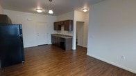 This single-floor space offers a clean, open-concept layout that seamlessly connects the kitchen and living areas, creating a sense of spaciousness and easy flow. The flooring throughout is rich hardwood-style planking in a warm brown tone, lending a grounded, cohesive feel to the interior.

The kitchen is positioned centrally within the open plan and is outfitted with dark espresso-toned cabinetry featuring ample storage above and below. A granite-style countertop runs along the kitchen base, paired with a stainless sink and black appliances including a dishwasher. Pendant lighting above the kitchen area adds a stylish focal point, complementing the flush-mount ceiling fixtures found throughout the space.

The walls are finished in a neutral light gray, providing a versatile backdrop that enhances the brightness of the unit. A doorway visible from the main living area leads into an additional room, offering further functional space. A paneled entry door provides access to the unit, contributing to the overall clean and well-maintained aesthetic.

The layout is efficient and thoughtfully designed, making strong use of its single-floor footprint to deliver a comfortable and functional living environment with a modern, understated character.
