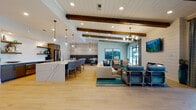 Located at 1079 Beckstead Ct in Denver, NC, this thoughtfully designed single-floor space offers an expansive open-plan layout that seamlessly integrates a gourmet kitchen, comfortable lounge, and stylish dining area. Light wood-look tile flooring flows throughout, unifying the space and enhancing its bright, airy atmosphere.

The kitchen serves as a central anchor, featuring a generously sized waterfall quartz island with bar seating, sleek gray cabinetry, open wood shelving for display, stainless steel appliances including a French door refrigerator, and globe pendant lights that add a warm, modern touch.

The lounge area is defined by exposed rustic wood ceiling beams set against a shiplap-style ceiling, a teal accent wall, and large sliding glass doors that invite abundant natural light and provide direct access to an outdoor patio area. The seating arrangement, anchored by a neutral sofa and coordinating accent chairs, creates a welcoming gathering space.

Adjacent to the kitchen, a dedicated dining nook features a bold channeled gray banquette, marble-top bistro tables, and industrial-style pendant lighting, offering an intimate setting within the larger open floor plan. The cohesive coastal-inspired color palette of blues, teals, and warm neutrals gives the entire space a polished yet relaxed ambiance.