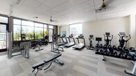 This fitness center is a thoughtfully designed facility that offers a comprehensive workout environment for all training needs. The space is anchored by large, floor-to-ceiling windows along one wall, flooding the room with abundant natural light and providing open views of the surrounding cityscape and greenery. A warm wood-paneled accent wall adds a refined architectural element, contrasting beautifully with the neutral tones of the patterned carpet tile flooring throughout.

The main workout floor is generously proportioned and well-organized into distinct training zones. The cardio section is equipped with multiple treadmills, elliptical machines, and Les Mills-branded stationary bikes, each featuring individual display screens. The strength training area includes a cable machine, a fully stocked dumbbell rack, a barbell station, and multiple adjustable benches. Free weights such as kettlebells and medicine balls are neatly stored along the windowed wall. Full-length mirrors span an entire wall, enhancing the sense of space while allowing users to monitor their form.

Ceiling fans provide additional ventilation and comfort throughout the space. An adjacent corridor features built-in white shelving units, a water fountain, and a patterned tile floor, serving as a transitional entry area leading to the main fitness floor. The overall ambiance is clean, modern, and energizing — a well-rounded facility designed to support a variety of fitness routines.