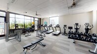 This fitness center is a thoughtfully designed facility that offers a comprehensive workout environment for all training needs. The space is anchored by large, floor-to-ceiling windows along one wall, flooding the room with abundant natural light and providing open views of the surrounding cityscape and greenery. A warm wood-paneled accent wall adds a refined architectural element, contrasting beautifully with the neutral tones of the patterned carpet tile flooring throughout.

The main workout floor is generously proportioned and well-organized into distinct training zones. The cardio section is equipped with multiple treadmills, elliptical machines, and Les Mills-branded stationary bikes, each featuring individual display screens. The strength training area includes a cable machine, a fully stocked dumbbell rack, a barbell station, and multiple adjustable benches. Free weights such as kettlebells and medicine balls are neatly stored along the windowed wall. Full-length mirrors span an entire wall, enhancing the sense of space while allowing users to monitor their form.

Ceiling fans provide additional ventilation and comfort throughout the space. An adjacent corridor features built-in white shelving units, a water fountain, and a patterned tile floor, serving as a transitional entry area leading to the main fitness floor. The overall ambiance is clean, modern, and energizing — a well-rounded facility designed to support a variety of fitness routines.